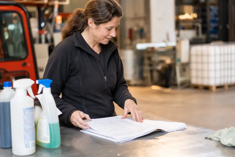 Betriebsleiterin prüft Sicherheitsdatenblätter und Reinigungsmittel auf einem Werkstatttisch.